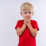 surprised-little-cute-blonde-boy-red-t-shirt-keeping-hands-his-mouth-while-looking-side-white-wall